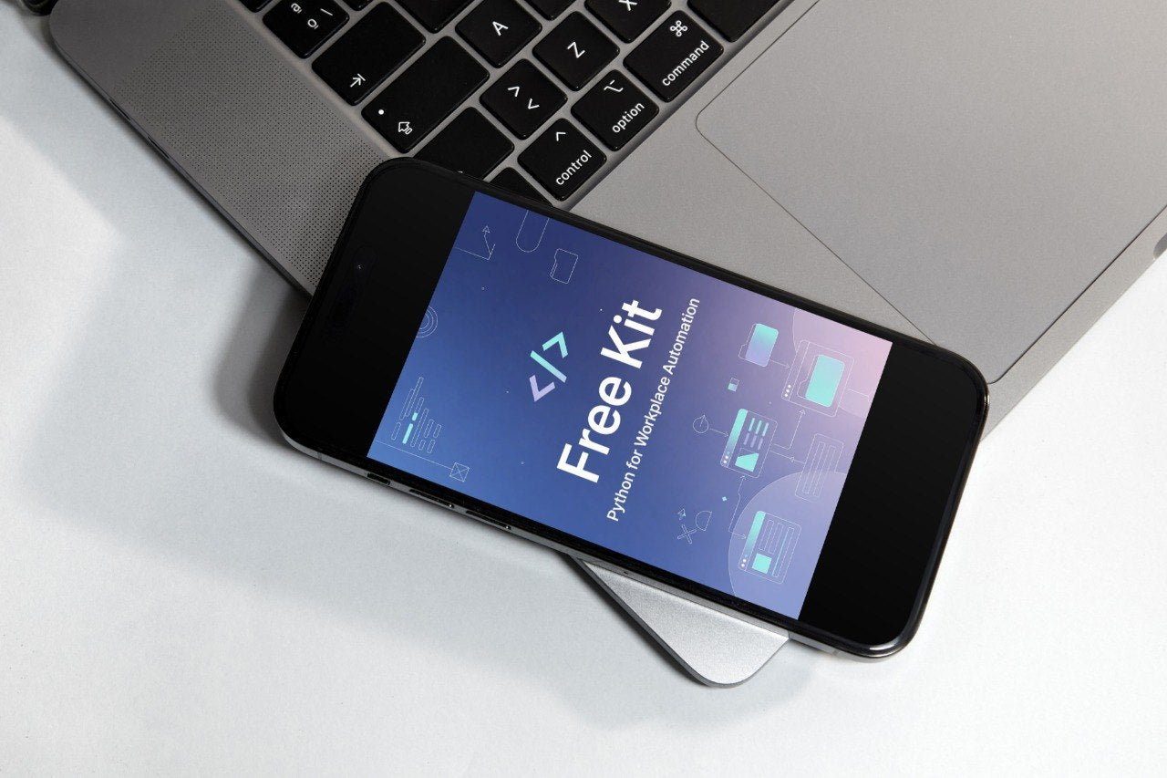 A black smartphone lying diagonally on a white surface, partially covering a silver laptop. The phone screen displays the text 'Free Kit' with a blue and purple gradient background.